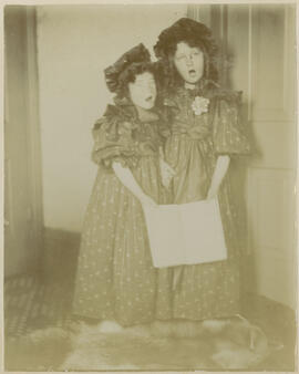 Two girls in matching dresses sing from a shared music sheet